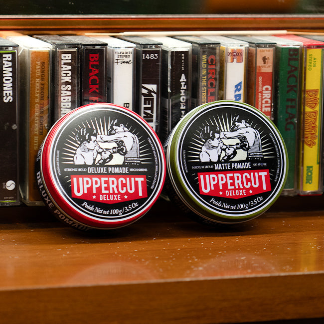 Uppercut Deluxe Matte Pomade and Deluxe Pomade tins leaning against cassettes